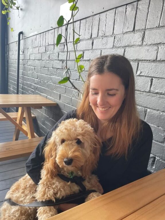 A woman sitting with a dog at picnic table, with brick wall and plants behind her, beginning her housesitting nomad life.
